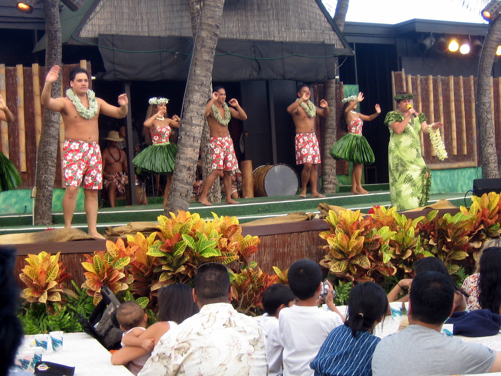 Dancers in grass skirts, board shorts, and leis perform on a stage.